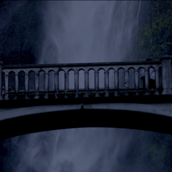 Stone bridge in front of a waterfall at dusk with surrounding foliage.
