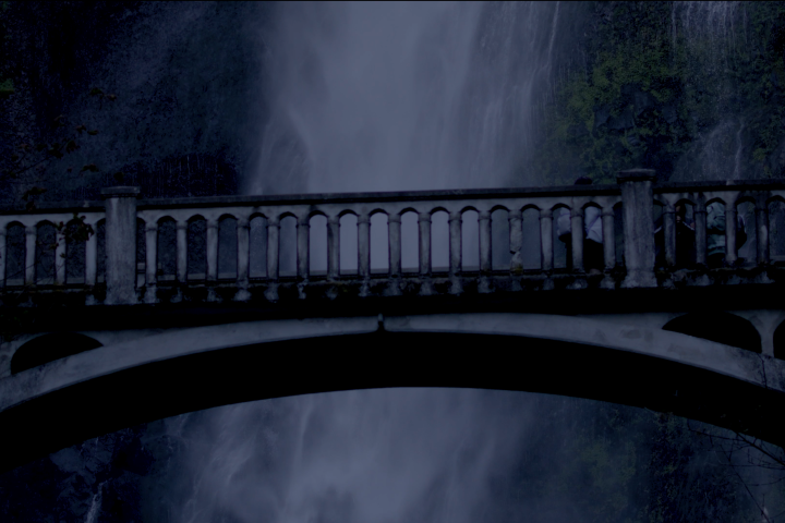 Stone bridge in front of a waterfall at dusk with surrounding foliage.