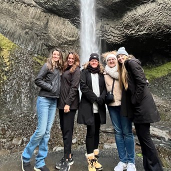 Five people posing together in front of a waterfall with rocky cliffs and mossy terrain.