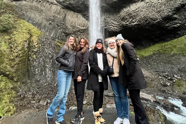 Five people posing together in front of a waterfall with rocky cliffs and mossy terrain.