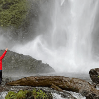 Person in red jacket stands with arms raised near a large waterfall.