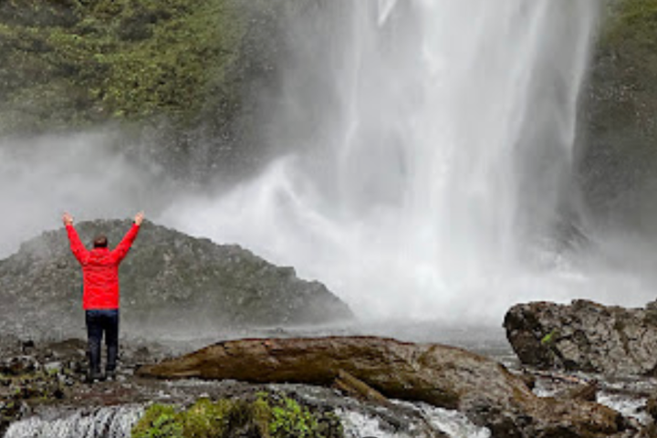 Person in red jacket stands with arms raised near a large waterfall.