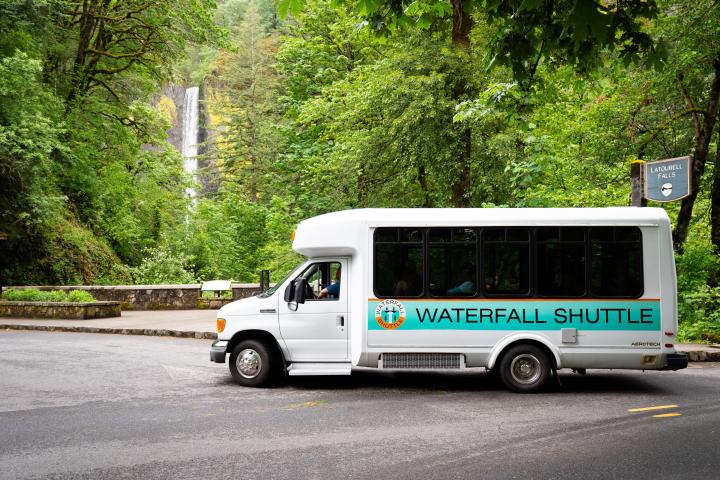 White shuttle bus parked near forest with waterfall and Latourell Falls sign.