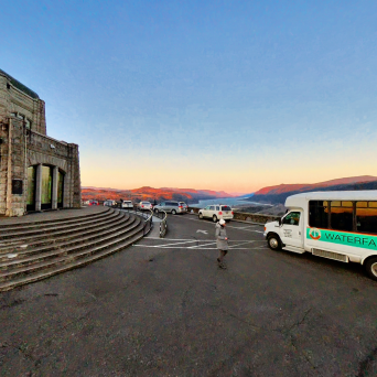 Stone building with stairs next to a shuttle bus in a parking area at sunset, overlooking a scenic landscape.