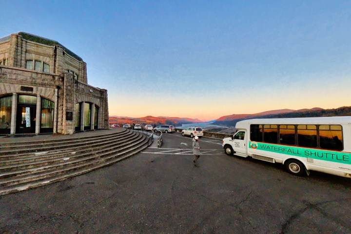 Stone building with stairs next to a shuttle bus in a parking area at sunset, overlooking a scenic landscape.