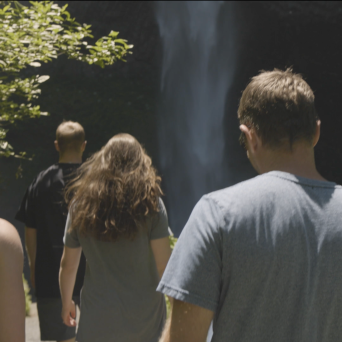 Four people walking towards a waterfall with greenery on the side.