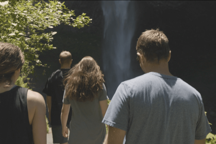 Four people walking towards a waterfall with greenery on the side.