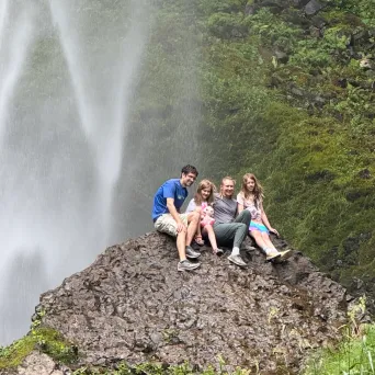 Group of people sitting on a rock near a waterfall in a green, mossy area.