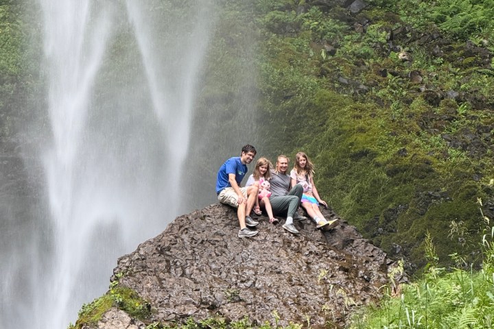 Group of people sitting on a rock near a waterfall in a green, mossy area.