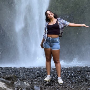 Person poses smiling in front of a waterfall with open arms, wearing casual clothing and sneakers.