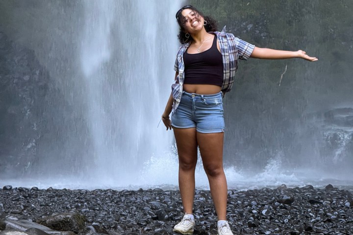 Person poses smiling in front of a waterfall with open arms, wearing casual clothing and sneakers.