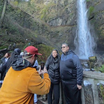 Tourists take photos by a waterfall in a forest setting, with a couple posing on a bridge.