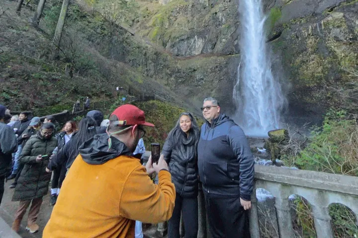 Tourists take photos by a waterfall in a forest setting, with a couple posing on a bridge.