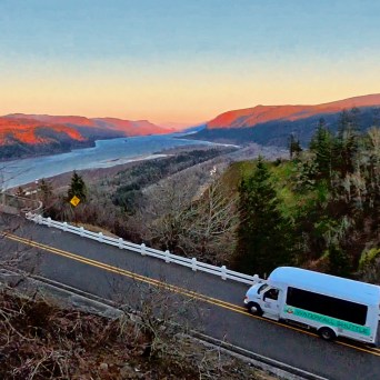 Bus on a winding road beside a river with hills during sunset.