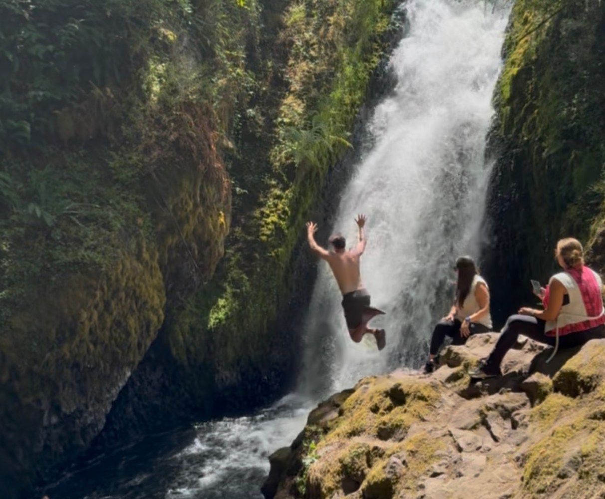Person jumping near waterfall with onlookers sitting on rocky surface.