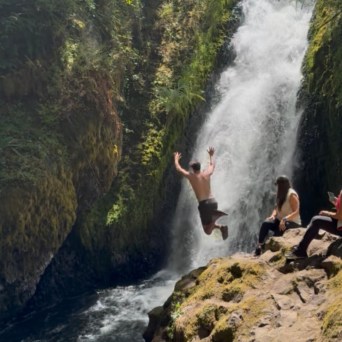 Person jumping near waterfall with onlookers sitting on rocky surface.