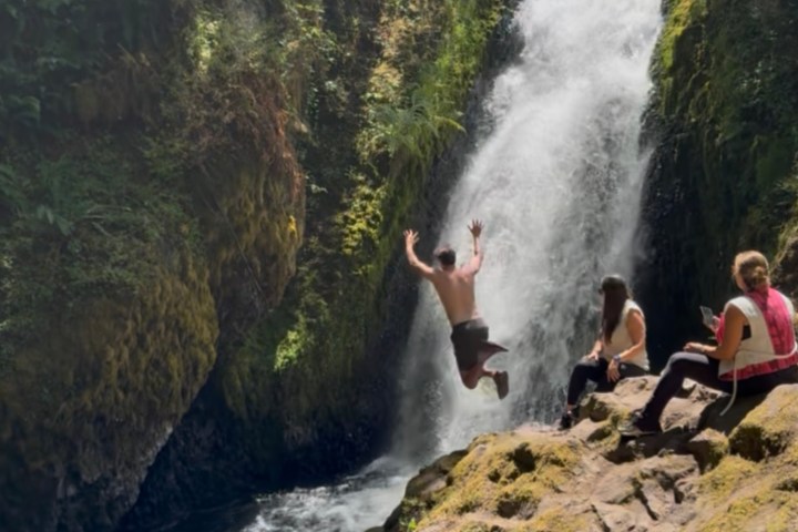 Person jumping near waterfall with onlookers sitting on rocky surface.