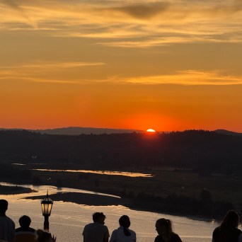 Silhouetted people watch a sunset over a river and distant hills.