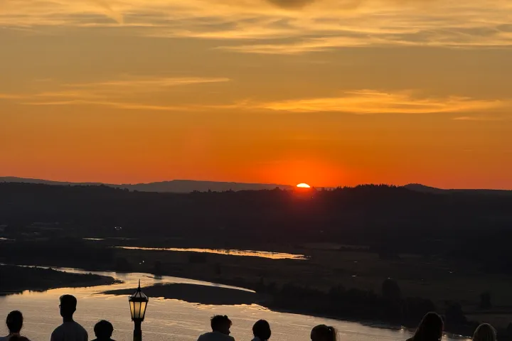 Silhouetted people watch a sunset over a river and distant hills.