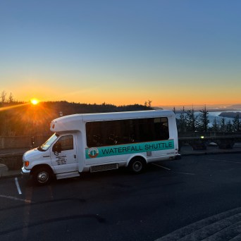 White shuttle bus parked at sunset with distant river and trees in background.