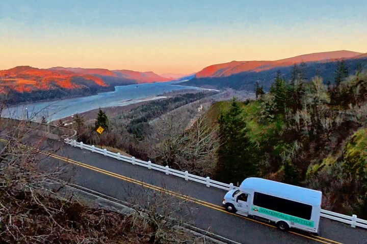 Bus on a winding road overlooking a river valley at sunset with mountains in the distance.