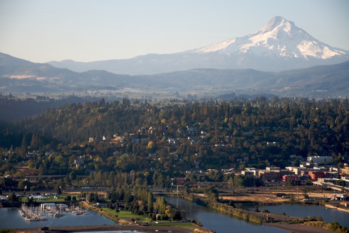 Scenic view of a town with a river and marina, forested hills, and a snow-capped mountain in the background.