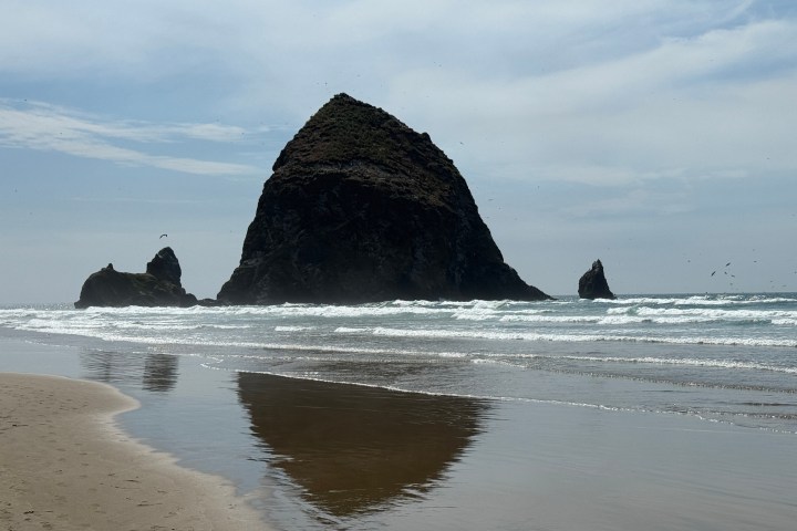 Large rock formation in ocean with waves and sandy beach under a cloudy sky.