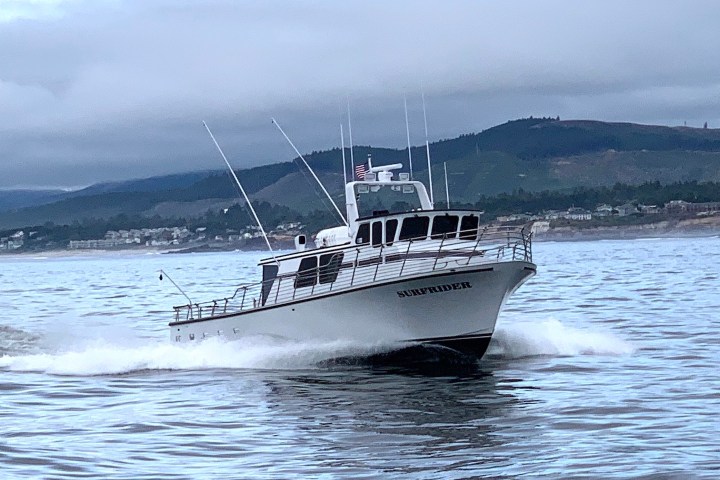 A white boat named Surfrider cruising on a calm sea with hills in the background.