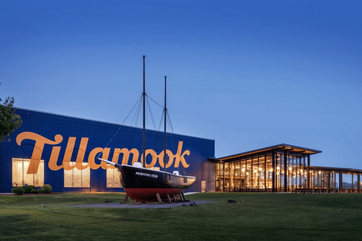 Building with 'Tillamook' logo, a boat on lawn, and modern architecture at dusk.