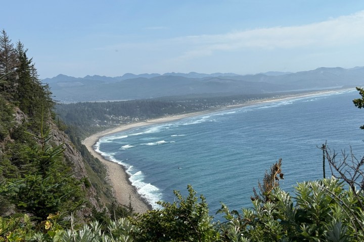 Coastal landscape with ocean waves, sandy beach, cliffs, and distant mountains under a clear sky.