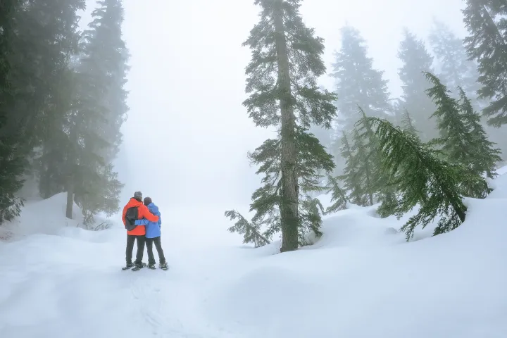 Two people in winter jackets stand in snowy mt hood forest with fog.